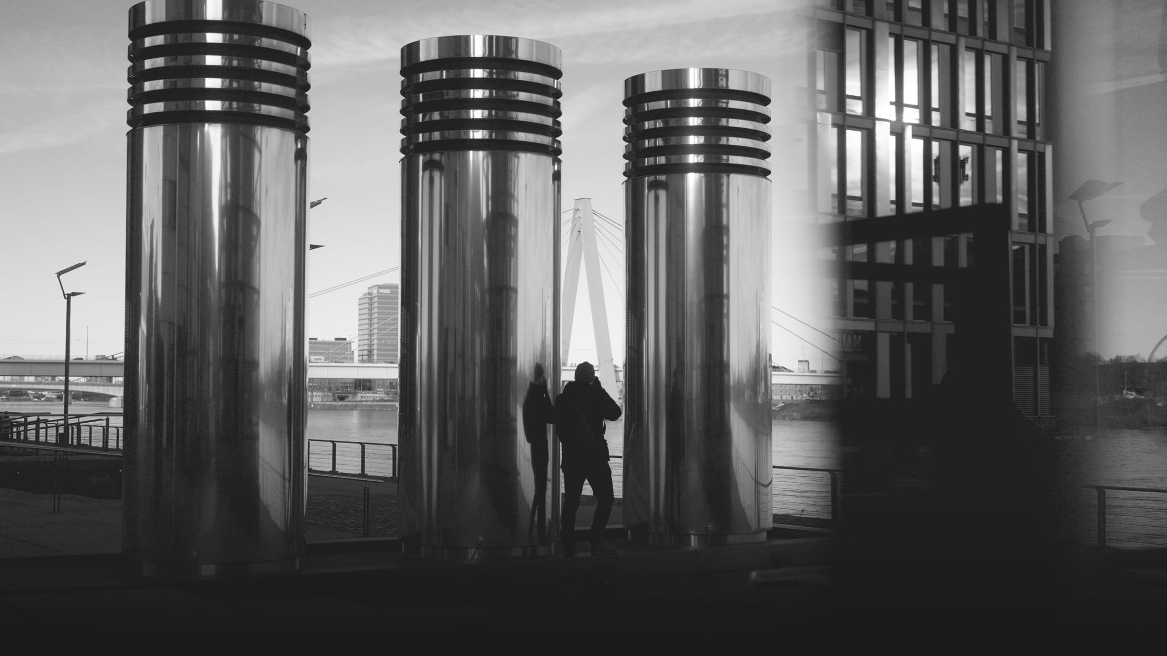 Photographer taking a picture between three large metallic cylinders in Cologne, near the Rheinauhafen. Silhouette reflection visible on the polished surfaces.