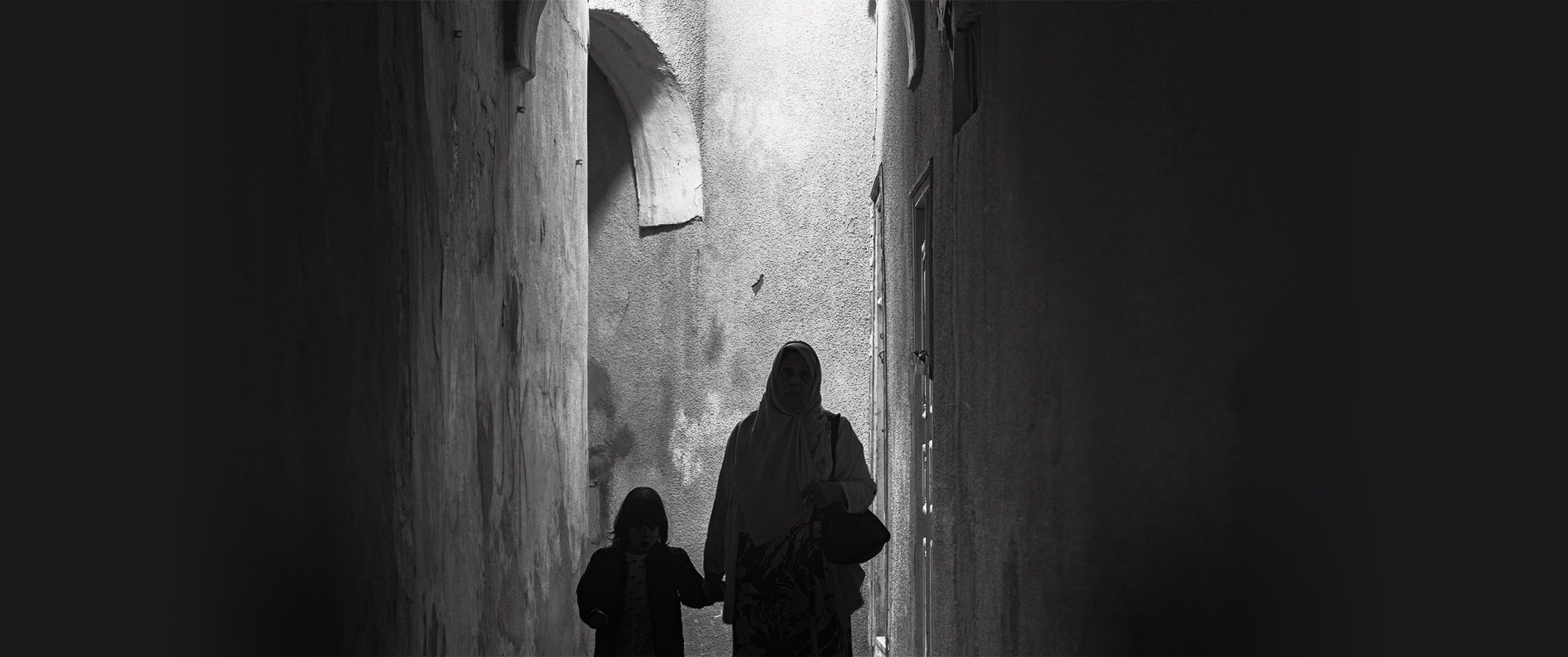 A woman and child walking through a narrow alley at night, illuminated by soft light. Monochrome photograph from Tunisia.