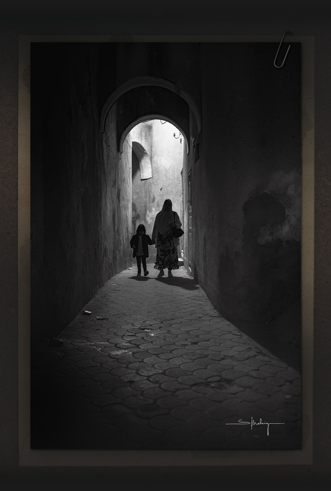 Black and white fine art photograph – mother and child walking through a narrow alley at night, illuminated by harsh artificial light