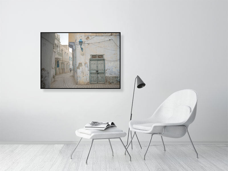 Photograph of a narrow alley with worn plaster walls, a faded green wooden door, and a blue street lamp in soft daylight. The muted tones and quiet street create a serene atmosphere reflecting the aged charm of Tunisian architecture.