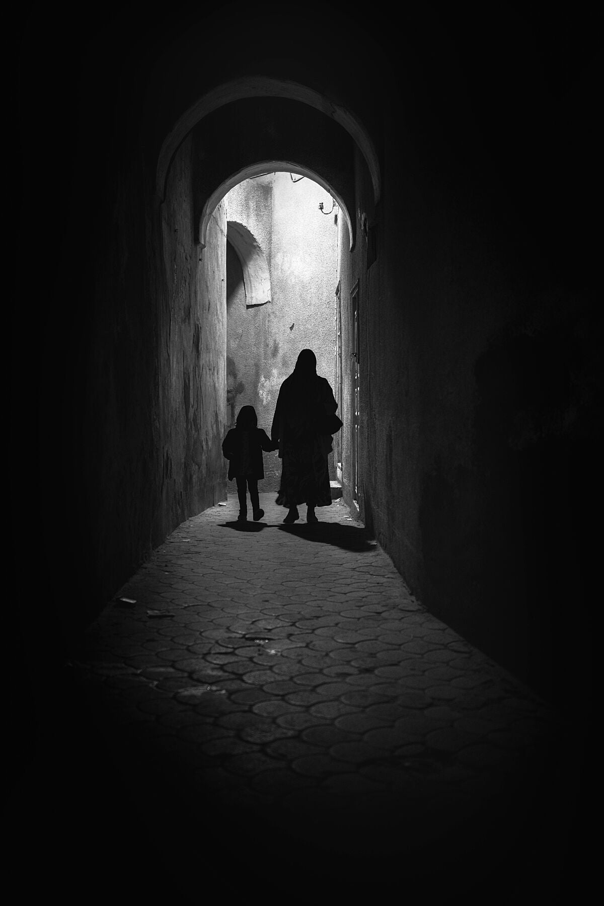 Monochrome photograph of a woman and child walking through a narrow alley under soft daylight. The scene evokes quiet observation and documentary calm — a defining image for the visual tone of Dustlight Archive.