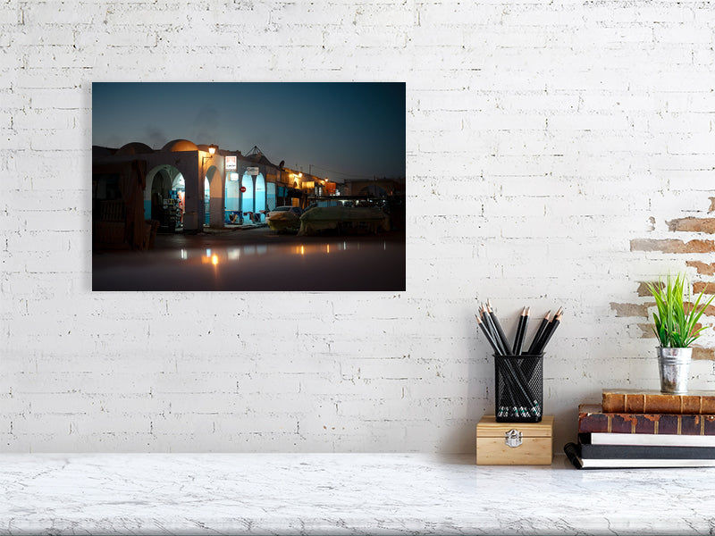 Evening photograph of a quiet market street illuminated by warm streetlights during the blue hour. Arched facades, parked cars, and covered stalls create a cinematic atmosphere, blending calm light with deep twilight tones.