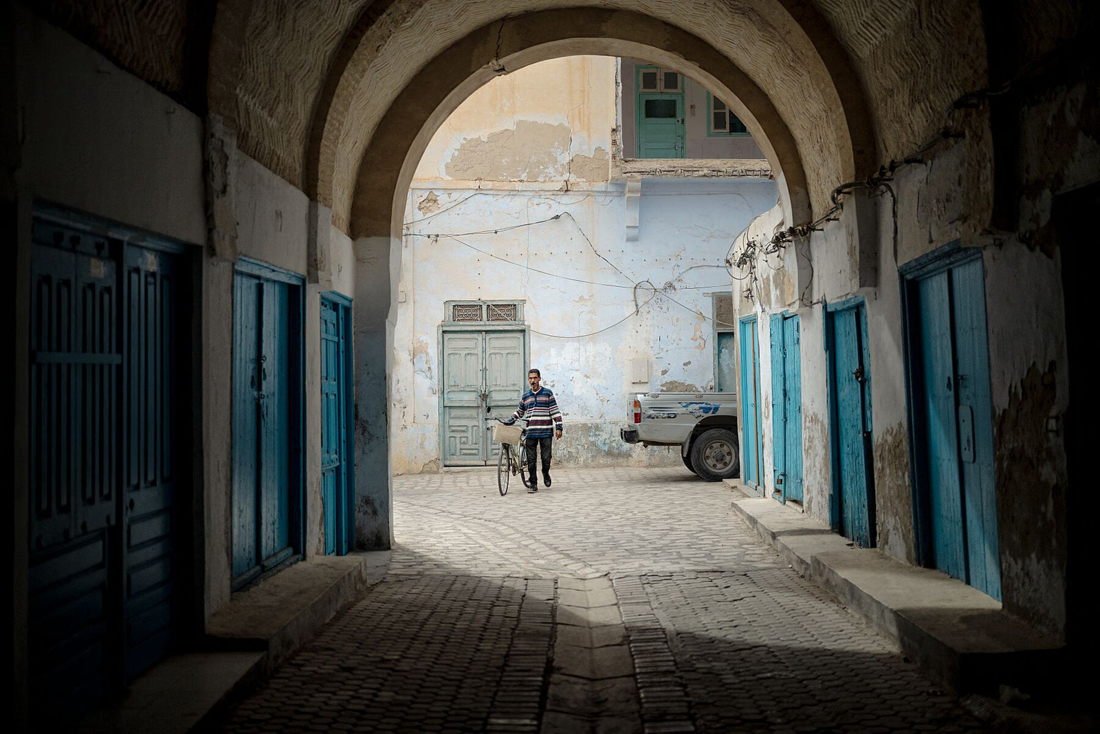 Photograph of a man walking a bicycle through an old stone archway into a sunlit alley with weathered blue doors and faded walls. The scene captures the quiet atmosphere of a Tunisian street, balancing shadow, light, and soft color tones.