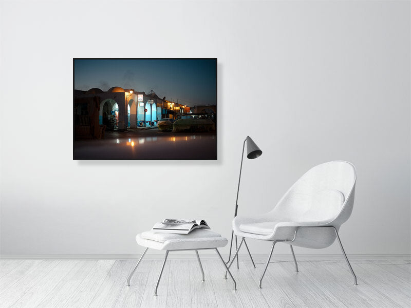 Evening photograph of a quiet market street illuminated by warm streetlights during the blue hour. Arched facades, parked cars, and covered stalls create a cinematic atmosphere, blending calm light with deep twilight tones.
