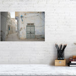Photograph of a narrow alley with worn plaster walls, a faded green wooden door, and a blue street lamp in soft daylight. The muted tones and quiet street create a serene atmosphere reflecting the aged charm of Tunisian architecture.