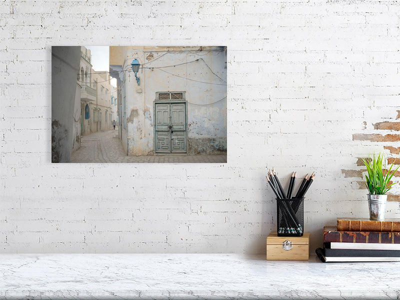Photograph of a narrow alley with worn plaster walls, a faded green wooden door, and a blue street lamp in soft daylight. The muted tones and quiet street create a serene atmosphere reflecting the aged charm of Tunisian architecture.