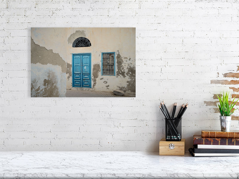 Photograph of a weathered building facade with a bright blue wooden door and matching window frame, both accented by ornate black metalwork. The peeling plaster and soft light reveal the quiet charm and patina of age in Tunisian architecture.