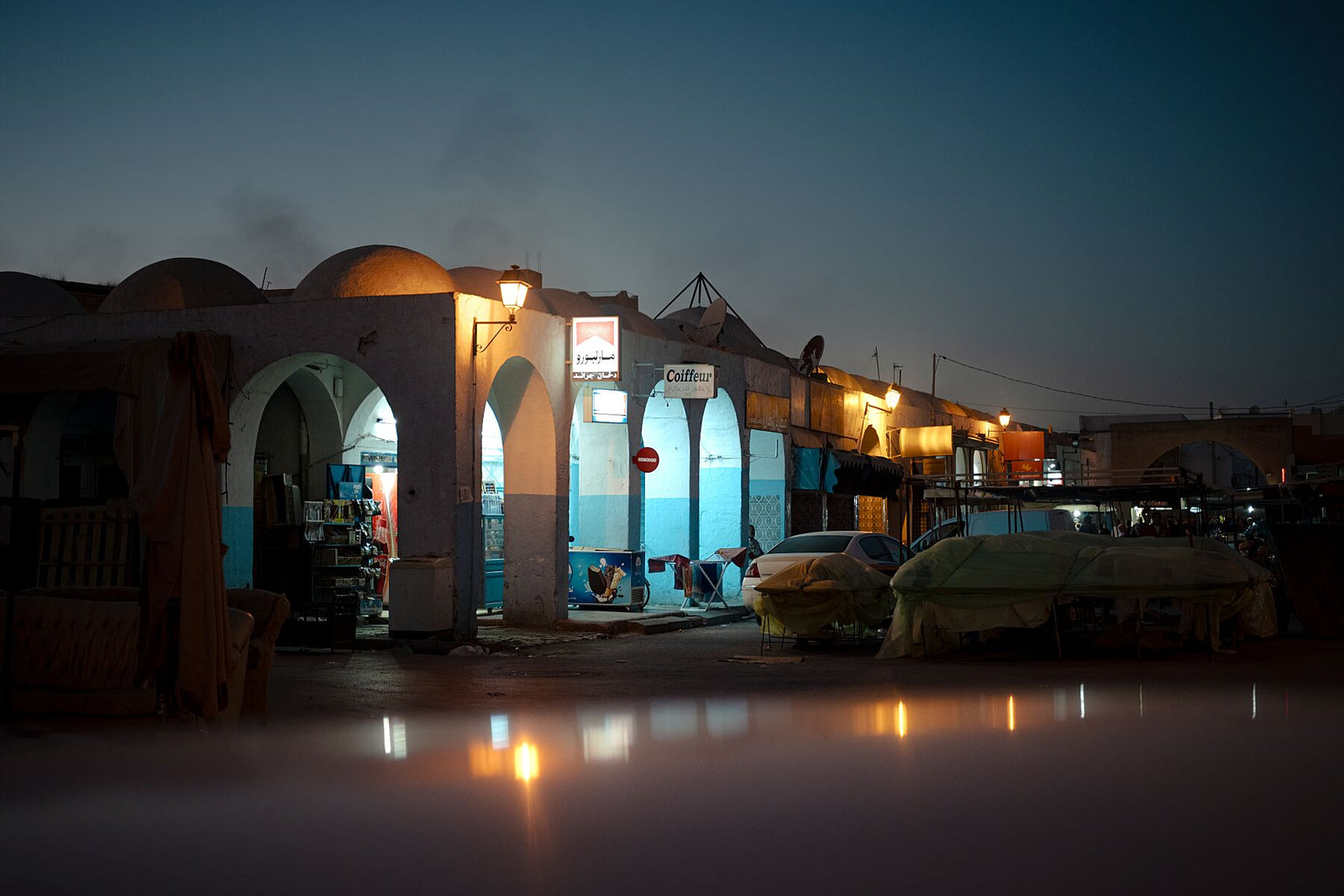 Evening photograph of a quiet market street illuminated by warm streetlights during the blue hour. Arched facades, parked cars, and covered stalls create a cinematic atmosphere, blending calm light with deep twilight tones.