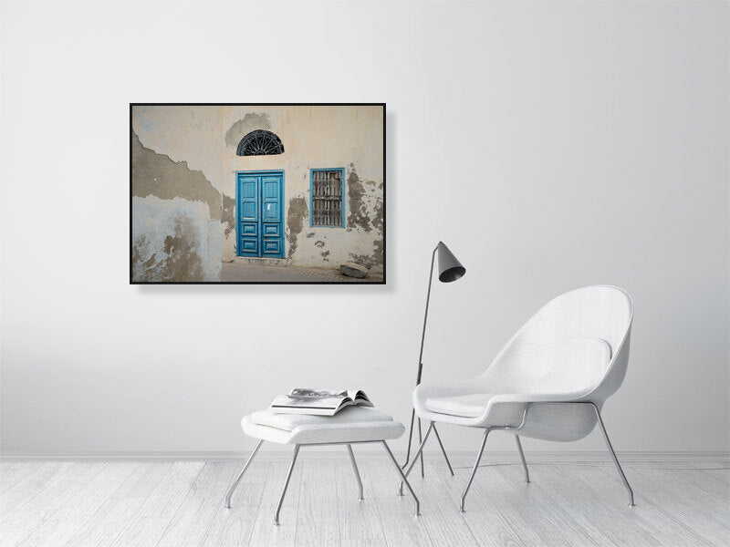 Photograph of a weathered building facade with a bright blue wooden door and matching window frame, both accented by ornate black metalwork. The peeling plaster and soft light reveal the quiet charm and patina of age in Tunisian architecture.