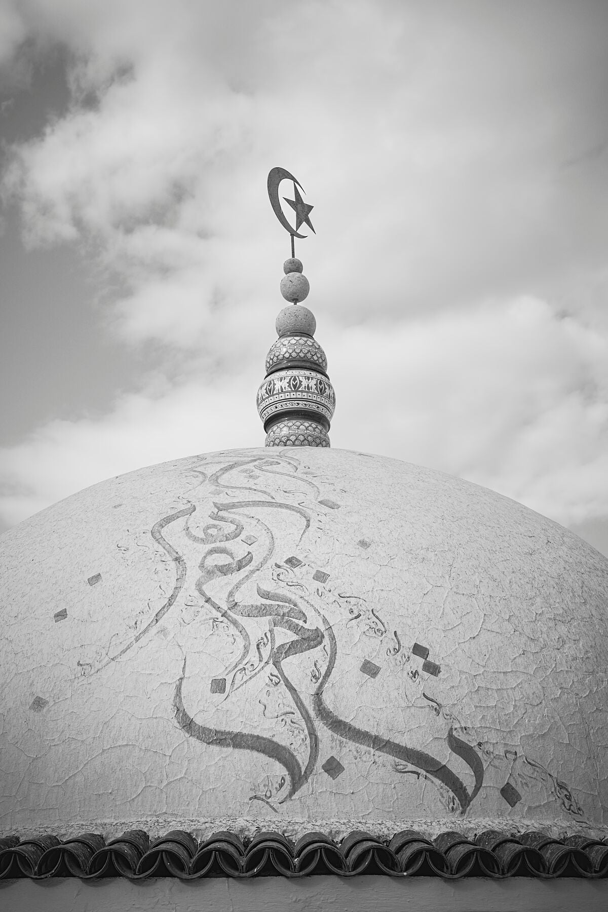Black-and-white photograph of a mosque dome with intricate Arabic calligraphy and a crescent moon and star finial, set against a soft cloudy sky. The image highlights texture, symmetry, and calm atmosphere in a fine art composition.