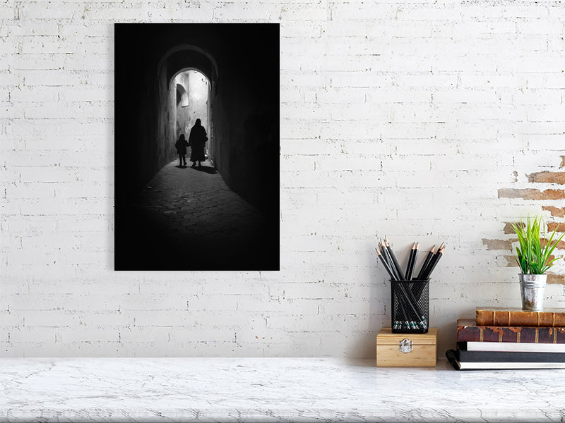 Monochrome photograph of a woman and child walking through a narrow alley under soft daylight. The scene evokes quiet observation and documentary calm — a defining image for the visual tone of Dustlight Archive.
