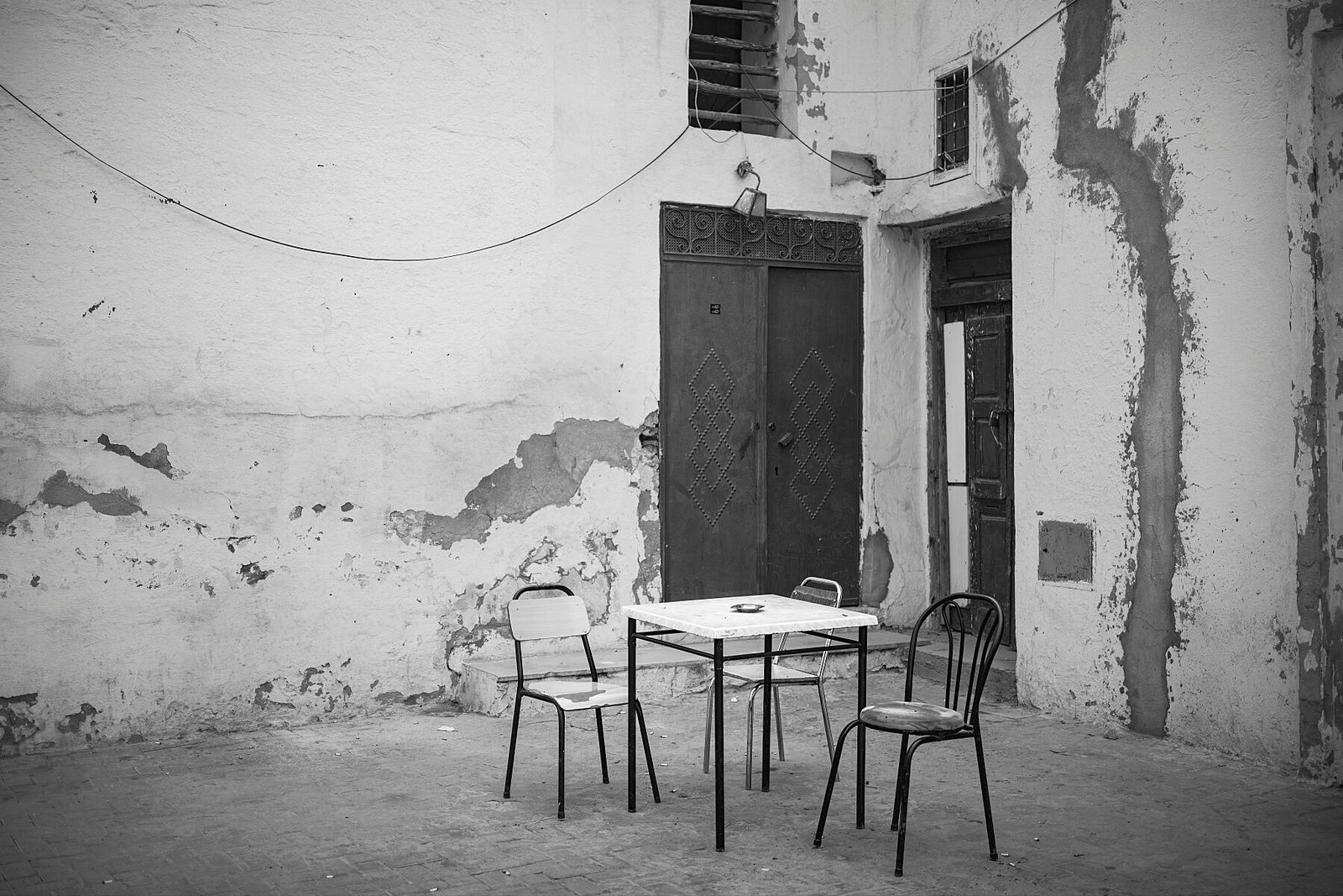Black and white photograph of three empty chairs around a small table set against a worn plaster wall with peeling paint and a dark metal door. The quiet courtyard scene conveys stillness, age, and a sense of absence.