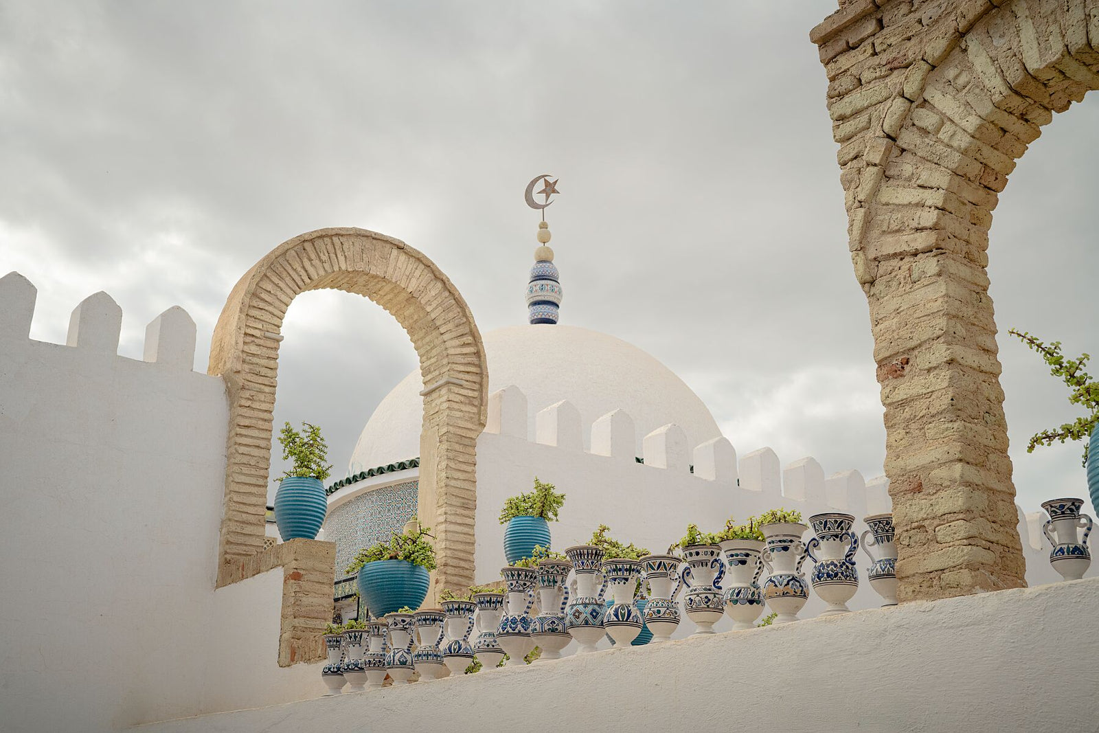 Photograph of a white dome with a crescent and star finial framed by stone arches and blue ceramic pots on a terrace in Tunisia. The scene captures calm Mediterranean architecture under a cloudy sky, emphasizing geometry, texture, and soft light.
