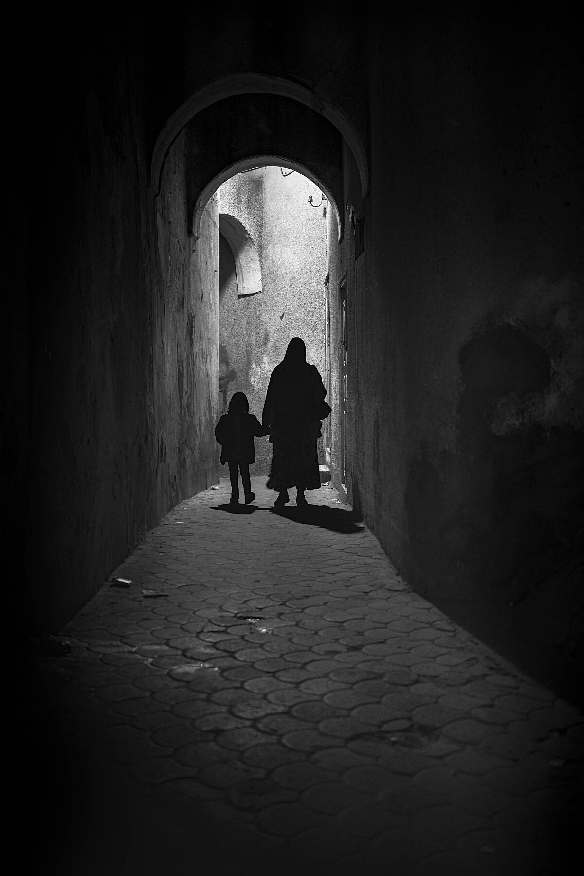 Black and white photograph of a narrow alleyway where a woman and a child walk hand in hand toward the light. The arched passage, textured walls, and deep shadows create a sense of intimacy, stillness, and timeless atmosphere.