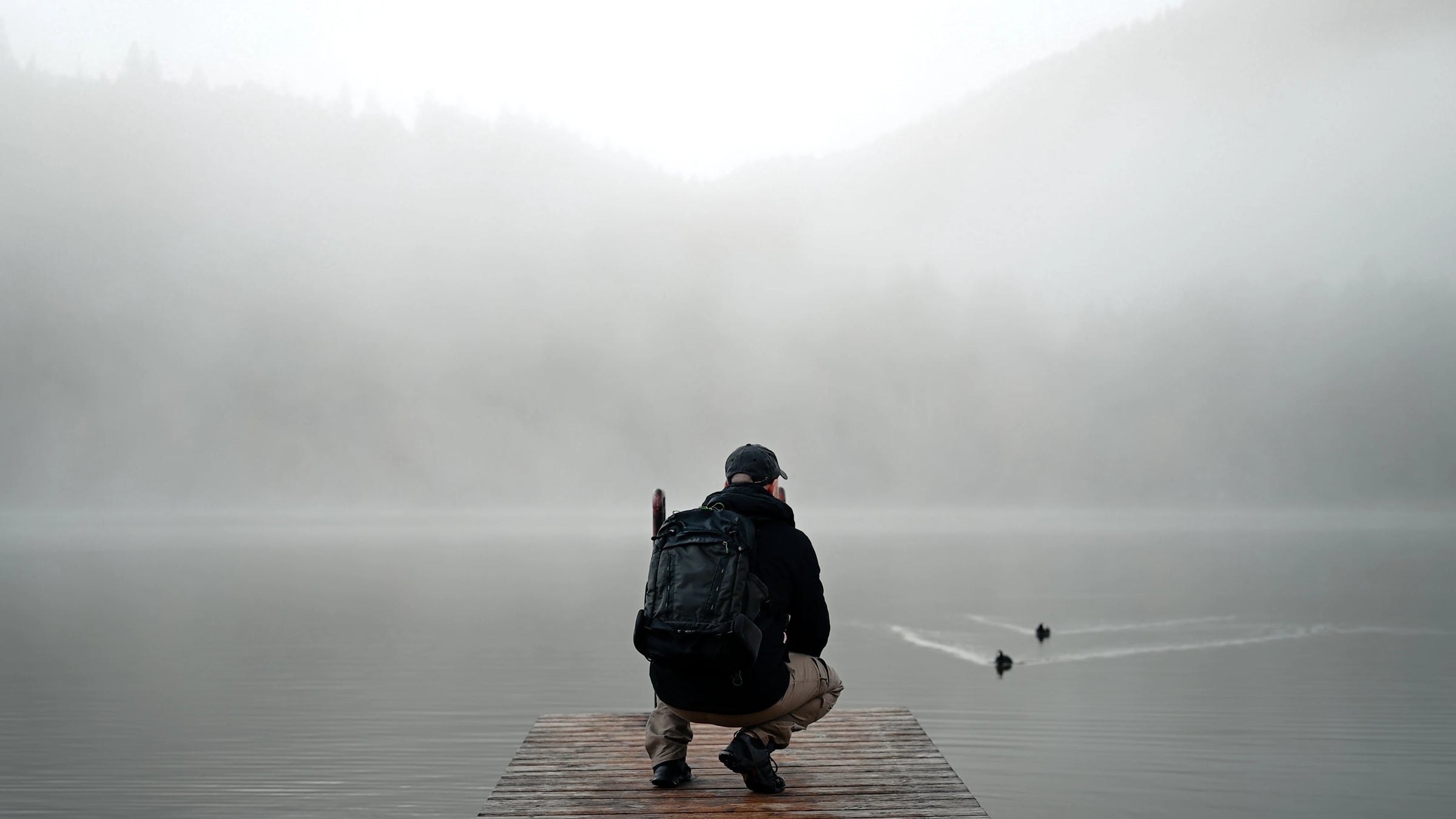A photographer crouches at the end of a fog-covered pier, quietly observing two ducks gliding across a still lake — a calm, cinematic moment from Through the Poetic Lens.
