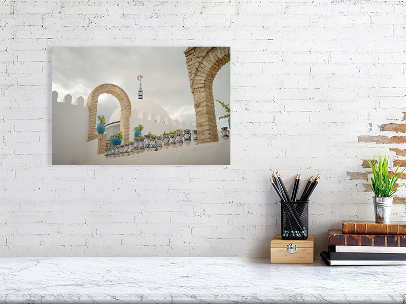Photograph of a white dome with a crescent and star finial framed by stone arches and blue ceramic pots on a terrace in Tunisia. The scene captures calm Mediterranean architecture under a cloudy sky, emphasizing geometry, texture, and soft light.