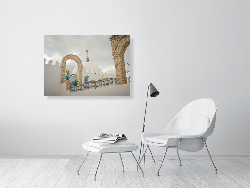 Photograph of a white dome with a crescent and star finial framed by stone arches and blue ceramic pots on a terrace in Tunisia. The scene captures calm Mediterranean architecture under a cloudy sky, emphasizing geometry, texture, and soft light.