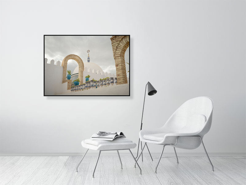 Photograph of a white dome with a crescent and star finial framed by stone arches and blue ceramic pots on a terrace in Tunisia. The scene captures calm Mediterranean architecture under a cloudy sky, emphasizing geometry, texture, and soft light.