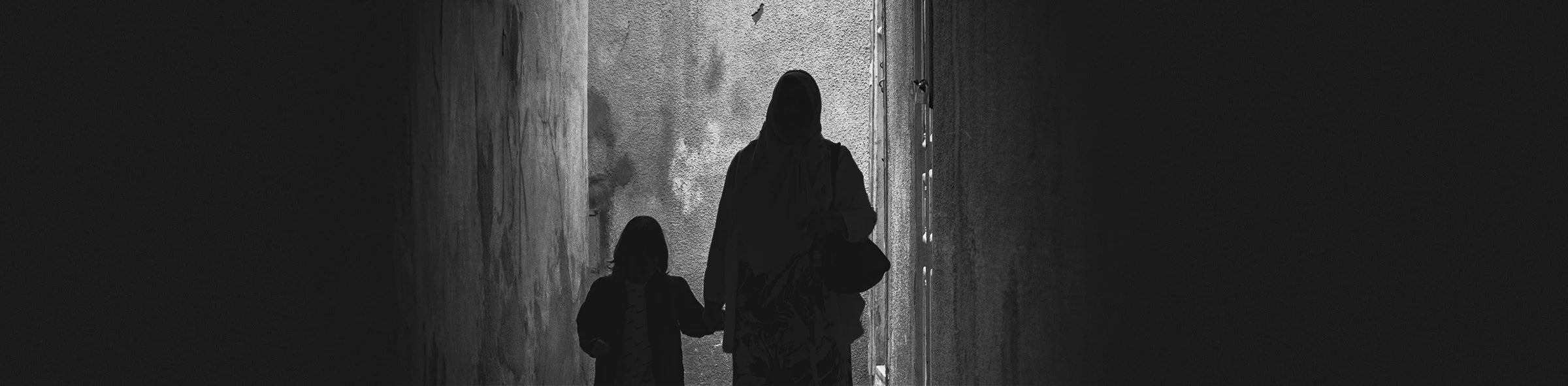 Monochrome photograph of a woman and child walking through a narrow alley, softly illuminated by ambient light at night. Scene captured in Tunisia.