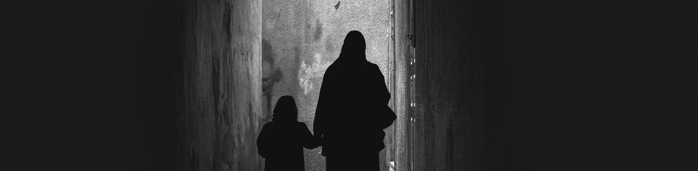 Monochrome photograph of a woman and child walking through a narrow alley, softly illuminated by ambient light at night. Scene captured in Tunisia.