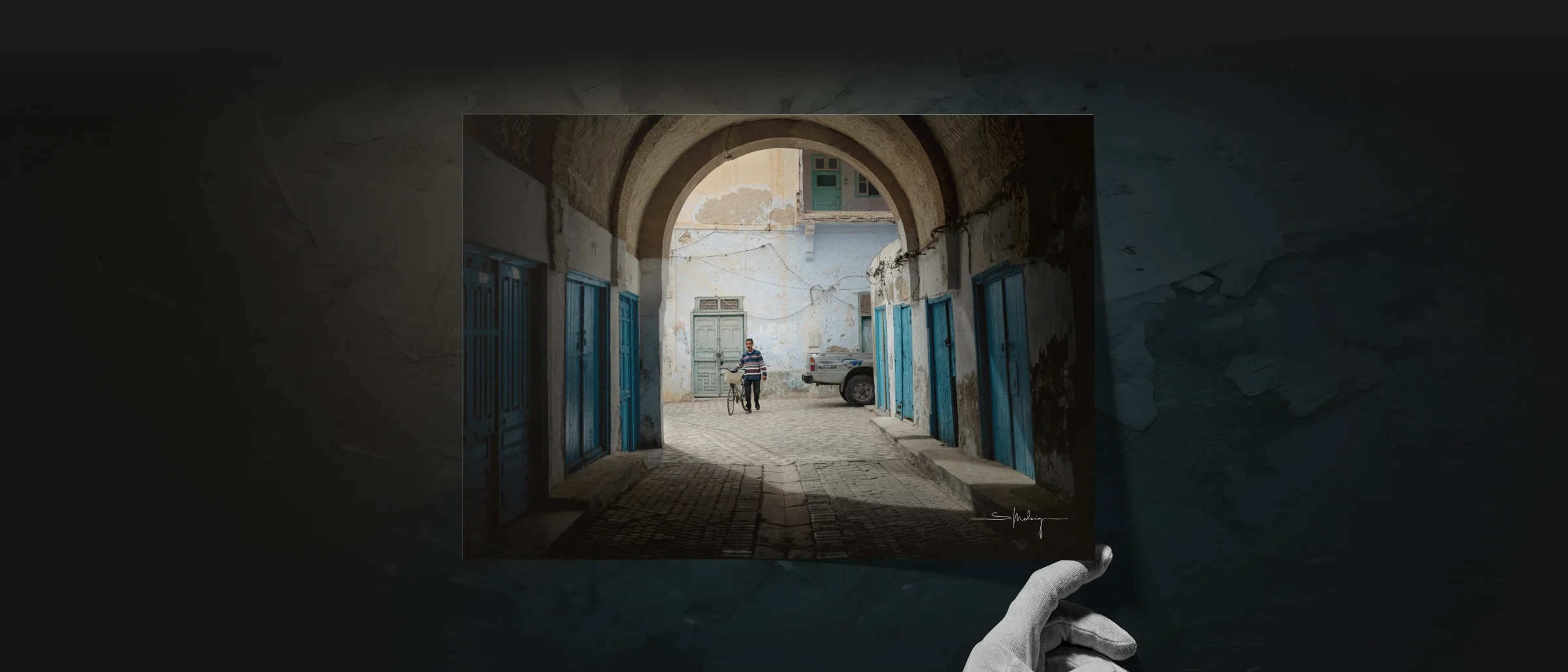 Photograph of a man walking his bicycle through an old archway framed by blue doors, shown as a print held by a sculpted hand. The scene bridges Dustlight’s cinematic storytelling with its fine art print collection.