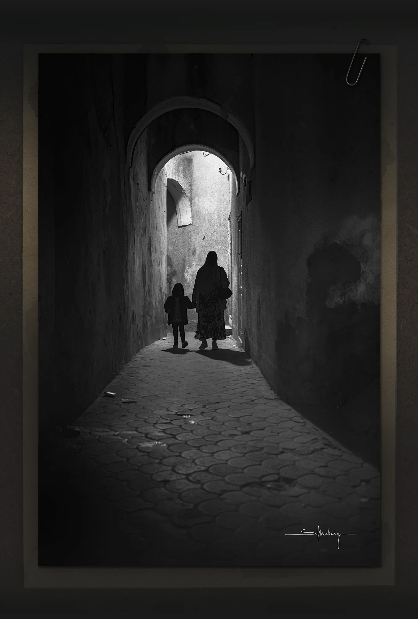 Monochrome photograph of a woman and child walking through a narrow alley under soft daylight. The scene evokes quiet observation and documentary calm — a defining image for the visual tone of Dustlight Archive.