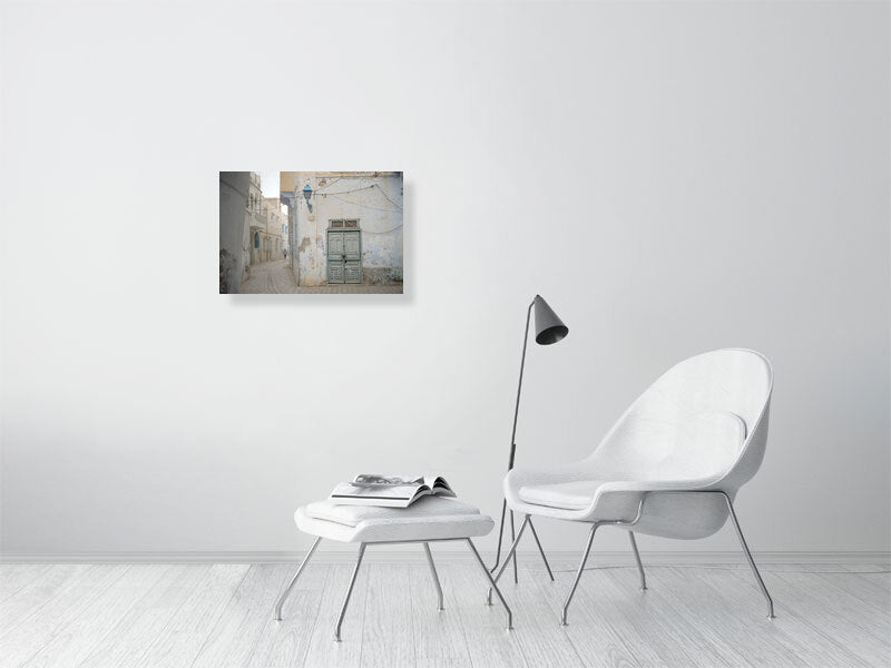Photograph of a narrow alley with worn plaster walls, a faded green wooden door, and a blue street lamp in soft daylight. The muted tones and quiet street create a serene atmosphere reflecting the aged charm of Tunisian architecture.