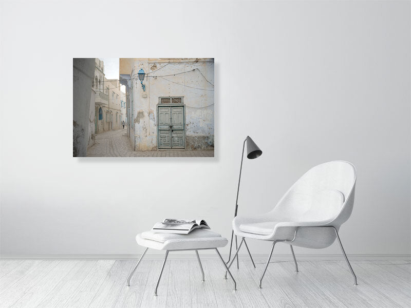 Photograph of a narrow alley with worn plaster walls, a faded green wooden door, and a blue street lamp in soft daylight. The muted tones and quiet street create a serene atmosphere reflecting the aged charm of Tunisian architecture.