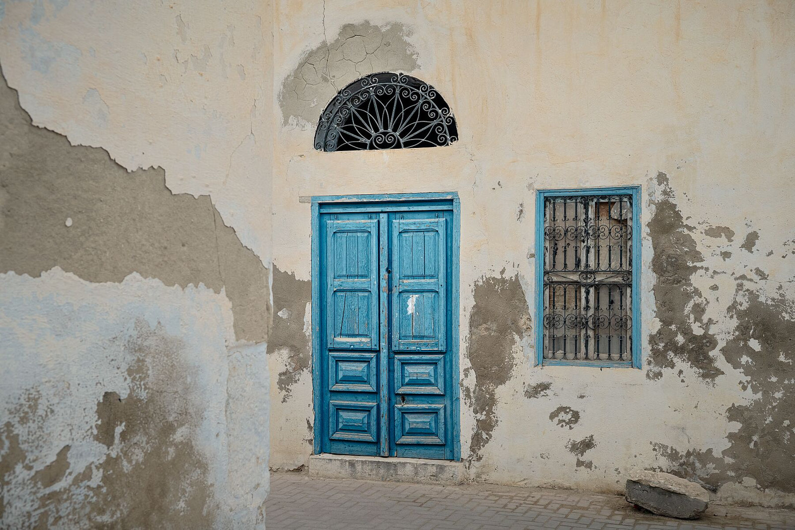 Photograph of a weathered building facade with a bright blue wooden door and matching window frame, both accented by ornate black metalwork. The peeling plaster and soft light reveal the quiet charm and patina of age in Tunisian architecture.