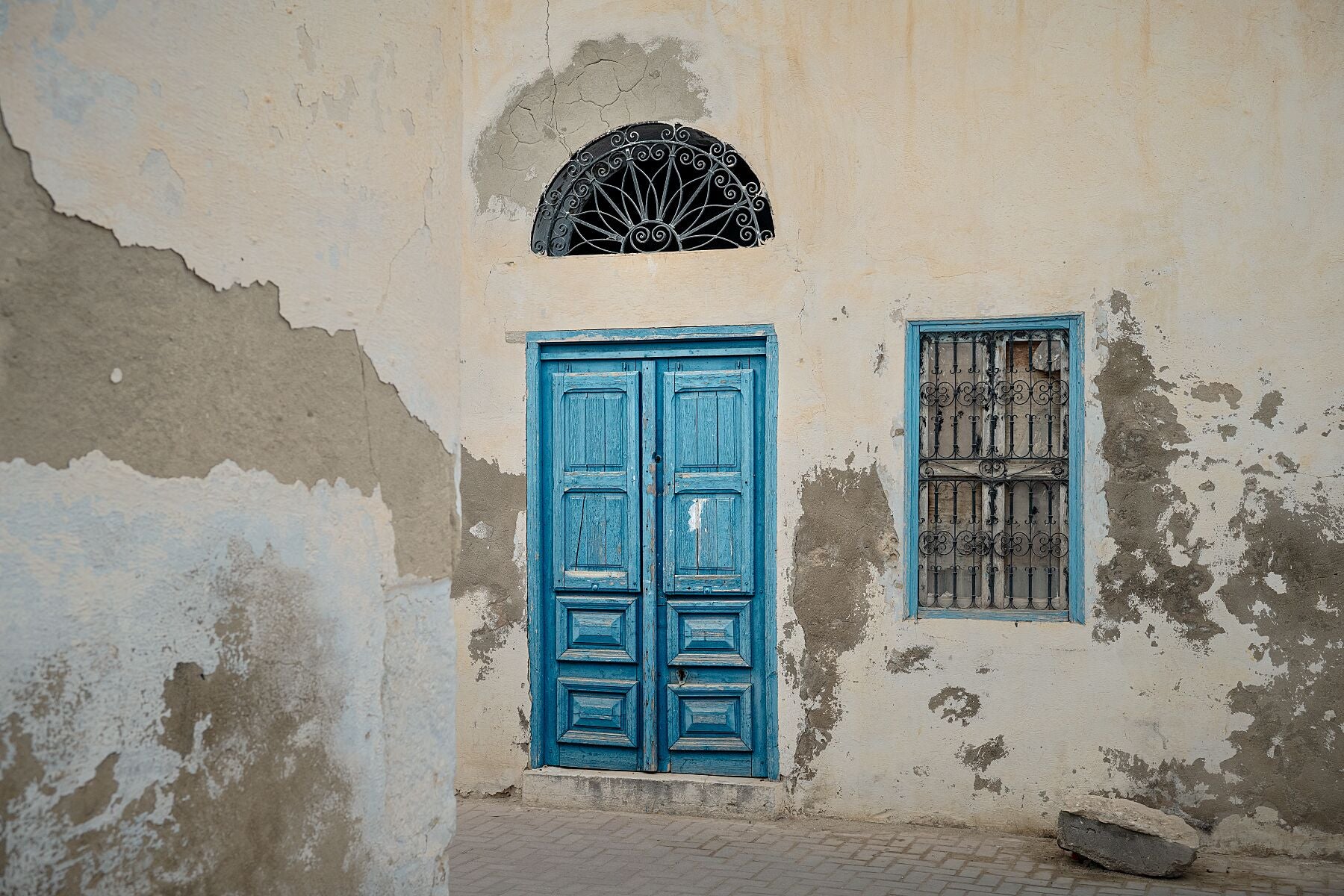Photograph of a weathered building facade with a bright blue wooden door and matching window frame, both accented by ornate black metalwork. The peeling plaster and soft light reveal the quiet charm and patina of age in Tunisian architecture.