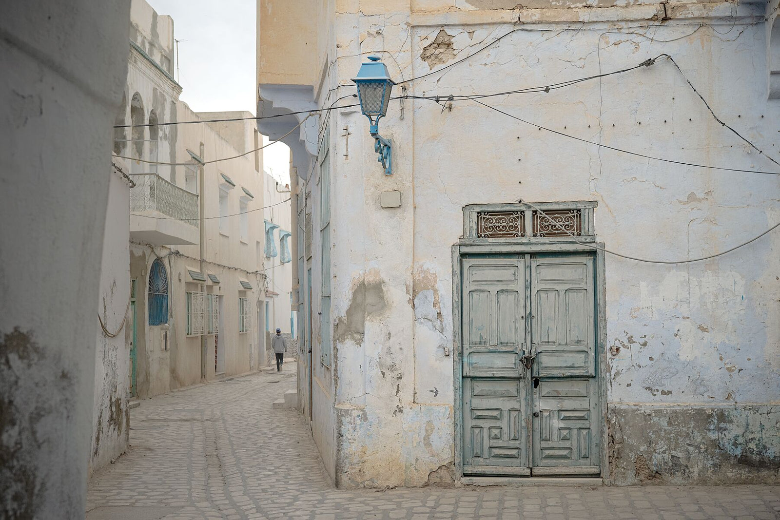 Photograph of a narrow alley with worn plaster walls, a faded green wooden door, and a blue street lamp in soft daylight. The muted tones and quiet street create a serene atmosphere reflecting the aged charm of Tunisian architecture.