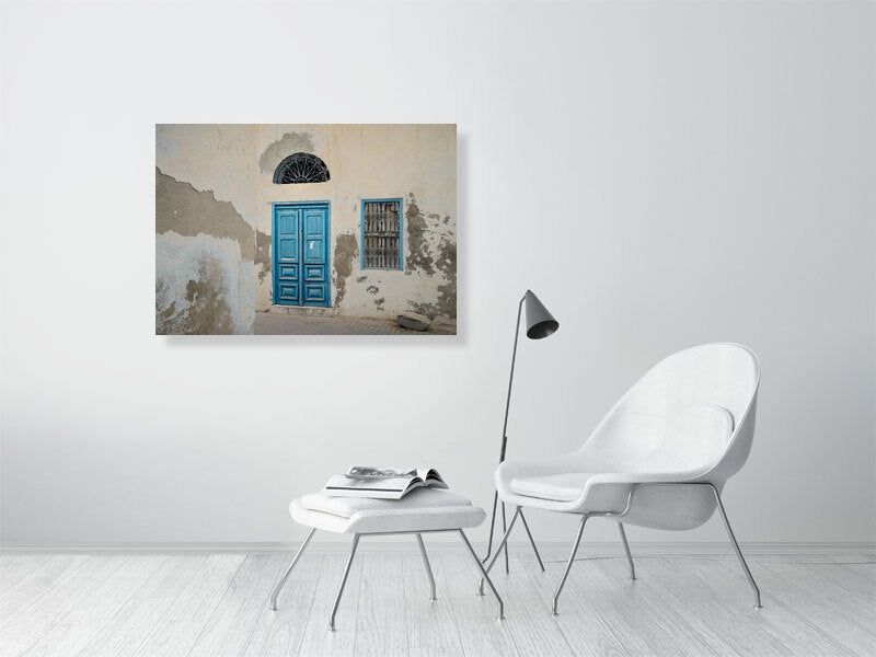 Photograph of a weathered building facade with a bright blue wooden door and matching window frame, both accented by ornate black metalwork. The peeling plaster and soft light reveal the quiet charm and patina of age in Tunisian architecture.